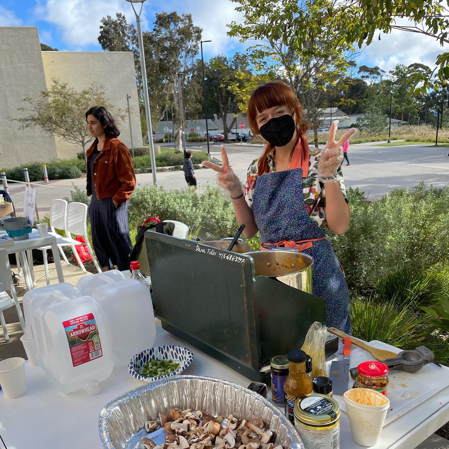 An outdoor area, partially paved and partially landscaped. A person, wearing a black face mask and an apron, makes two 'peace' signs with their plastic-gloved hands. In front of them is a gas camp stove, some jugs of water, a pan of mushrooms, and an assortment of jars. A person stands in the background, looking to the side.