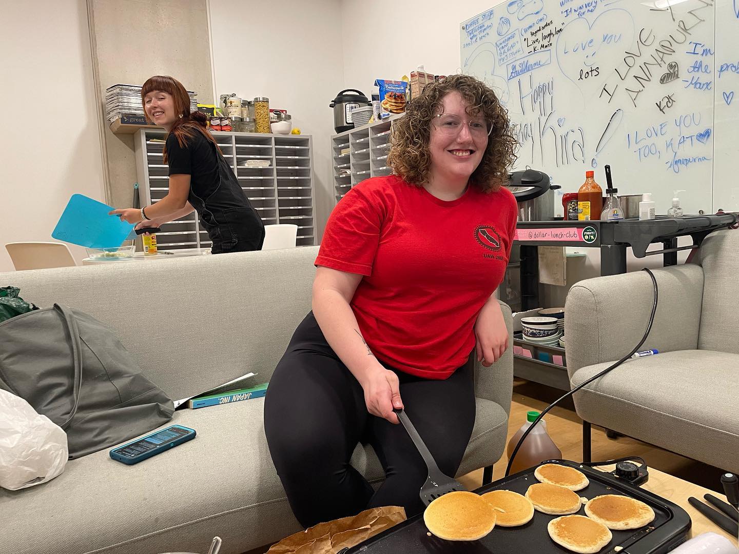 A person sitting on a couch smiles at the camera while they flip pancakes on an electric griddle. A person in the background smiles at the camera while holding a cutting mat.